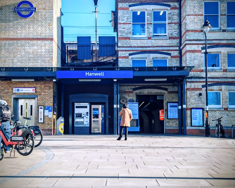 A person standing outside the entrance to Hanwell station, part of the Elizabeth line, with a woman in a beige coat and jeans facing the ticket machines and information boards mounted on the wall. The station building is constructed from brick with red and cream tones, featuring large windows on the upper floors and a small balcony with metal railings. To the left, there are several bicycles parked, including one with a red and black design and branding, and a small parking area for bikes and scooters. The station entrance has a black canopy with a blue sign displaying the station name 'Hanwell', and next to the ticket machines, there is a small mailbox. The scene is illuminated by natural daylight, with a clear sky visible at the top of the image, and a vintage-style street lamp is mounted on the right side of the building. The context suggests a quiet moment in a residential or suburban area, with nearby facilities for commuters. This setting is related to home relocation or moving services supplied by Elephant and Castle Movers, as part of professional removals or furniture transport during house moves.