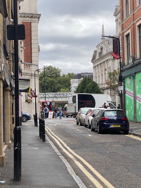 A street scene near Elephant and Castle with parked cars along the curb, including a black hatchback in the foreground, and a large white moving truck with branding parked on the right side, partially blocking the road. Multiple cardboard boxes and wrapped furniture pieces are visible being loaded onto the truck, with several people assisting in the home relocation process. The street is lined with historic brick and stone buildings, some with decorative architectural details, and a green construction barrier on the right. In the background, a bridge spans over the street, and pedestrians are walking on the sidewalk. Overcast sky above creates diffuse lighting, typical of a busy urban scene involving furniture transport and packing activities, with Elephant and Castle Movers conducting a professional removals service aligned with house moving and logistics.