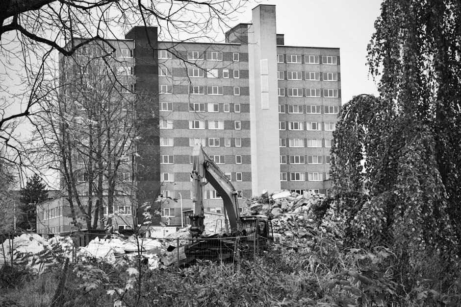 A construction site in an urban residential area featuring a multi-storey apartment building with numerous windows in the background. In the foreground, there is an excavator with a hydraulic arm positioned over a large pile of rubble and debris, including broken concrete, bricks, and rocks, indicating demolition or site clearance. Surrounding the uneven ground are patches of grass and small bushes, with leafless trees partly framing the scene. The scene is captured in black and white, with overcast lighting creating a neutral tone. This setting relates to the process of home relocation or renovation, where building demolition or site preparation is part of the moving or refurbishment process. Elephant and Castle Movers may use such sites during furniture transport or packing and moving operations involving clearance or access preparation for house removals.
