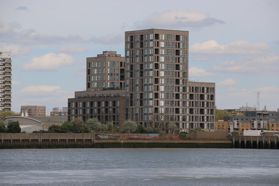 The image shows a modern multi-storey residential building located along a waterway with a reflective surface. The building features a contemporary design with a dark brick exterior and numerous large rectangular windows, arranged in an irregular pattern across its multiple sections, which vary in height and shape. The surrounding environment includes a mix of trees and lower-rise buildings, with some industrial or commercial structures visible nearby. The sky above is partly cloudy with patches of blue. In the foreground, a wooden barrier lines the water’s edge, suggesting a quay or riverside setting. The scene appears to be during daytime with natural lighting illuminating the building and its surroundings. This setting could be part of an urban relocation or home removal project managed by Elephant and Castle Movers, who specialise in furniture transport and packing during house removals in the Elephant Park area.