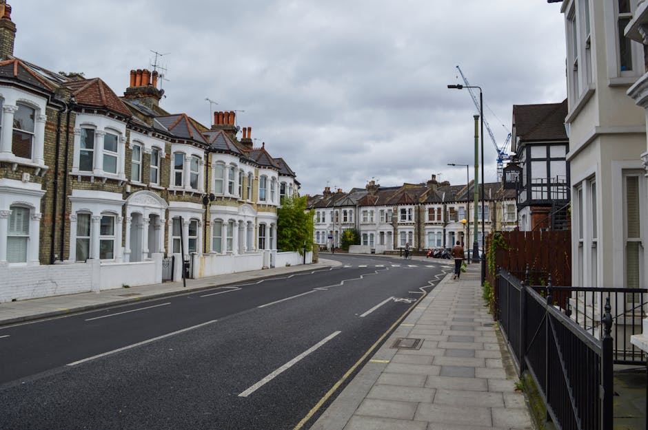 A residential street with Victorian-style terraced houses featuring bay windows, decorative brickwork, and chimneys, lined along a paved sidewalk with black metal railings. The street is empty except for a person walking further down the pavement. Overhead, the sky is overcast with grey clouds, and there are streetlights and a construction crane visible in the background. The scene appears calm, reflecting typical urban surroundings where house removals or moving services by Elephant and Castle Movers may take place, especially given the accessible street environment suited for furniture transport and loading processes.