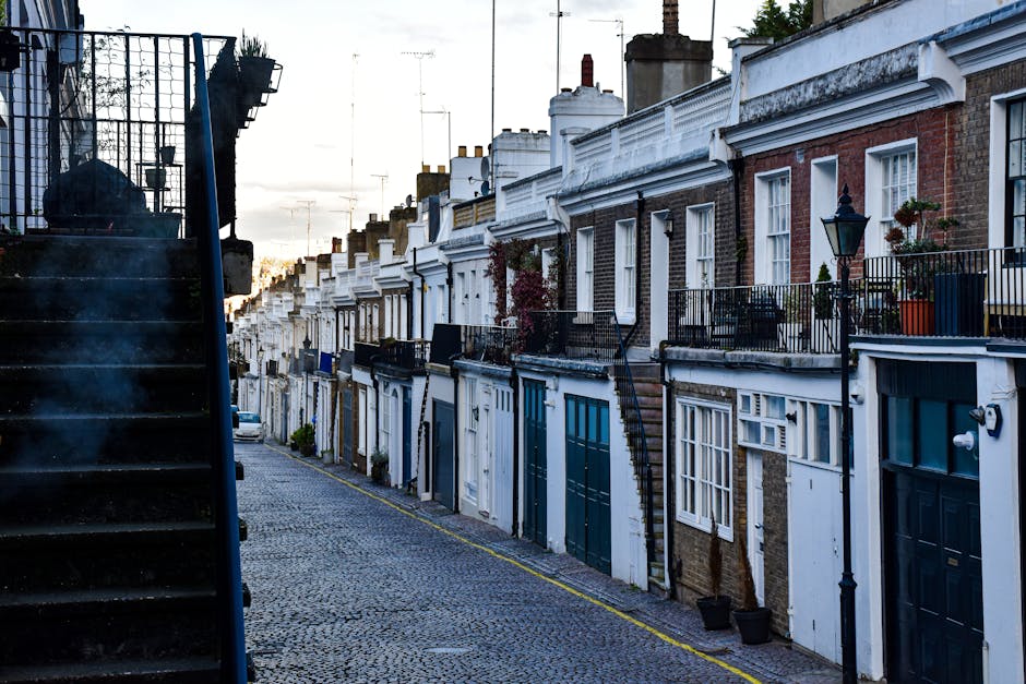 A narrow residential street on Walworth Road featuring a row of terraced houses with brick facades, some painted white and others with exposed brickwork. Each house has a small front garden or porch area, with potted plants and decorative railings. On the left side of the image, a dark exterior staircase with metal railing leads up to an upstairs entrance, casting shadows on the cobblestone pavement below. The street is lined with black iron street lamps and small trees in pots, adding to the neighborhood’s character. The cobbled road is empty, extending into the distance under a sky with a subtle sunset glow. This scene provides a typical backdrop for house removals and furniture transport, with the exterior environment ready for a home relocation process, as handled by Elephant and Castle Movers, supporting stair access and urban moving logistics.